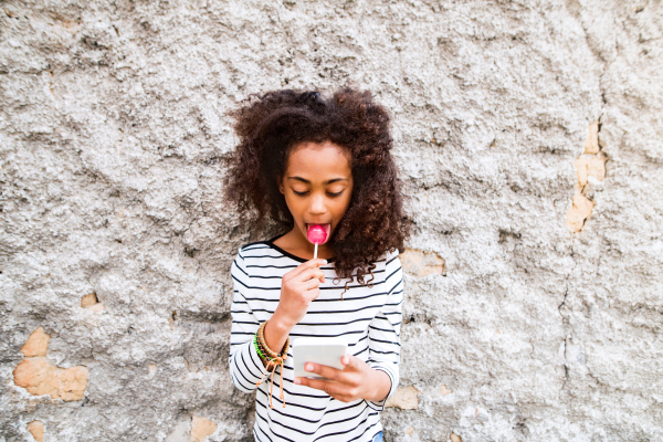 Beautiful african american girl with curly hair outdoors against concrete wall, holding smart phone, texting and eating lollipop.