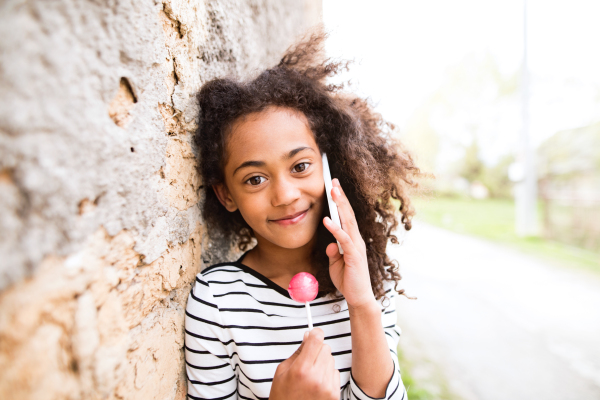 Beautiful african american girl with curly hair outdoors against concrete wall, holding smart phone, making phone call and eating lollipop.