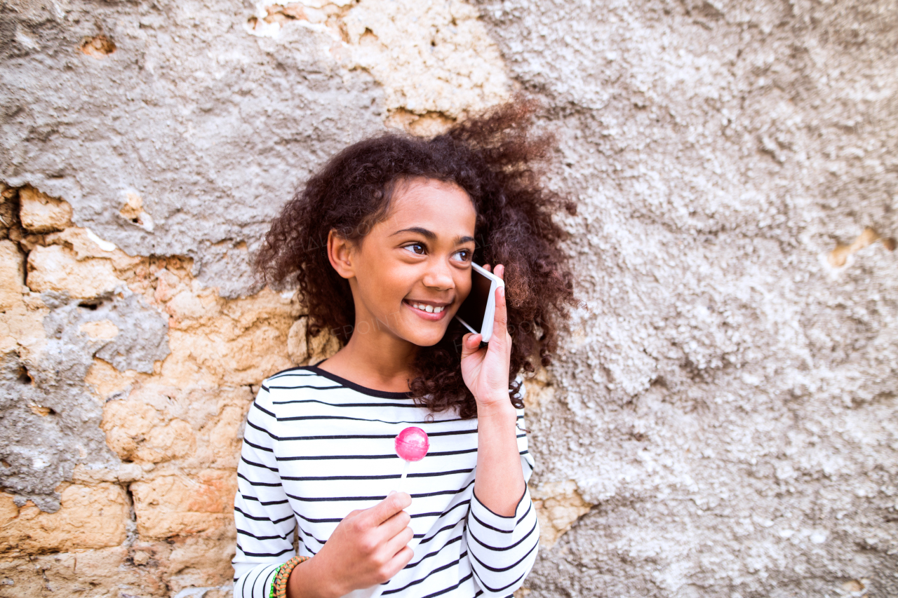 Beautiful african american girl with curly hair outdoors against concrete wall, holding smart phone, making phone call and eating lollipop.