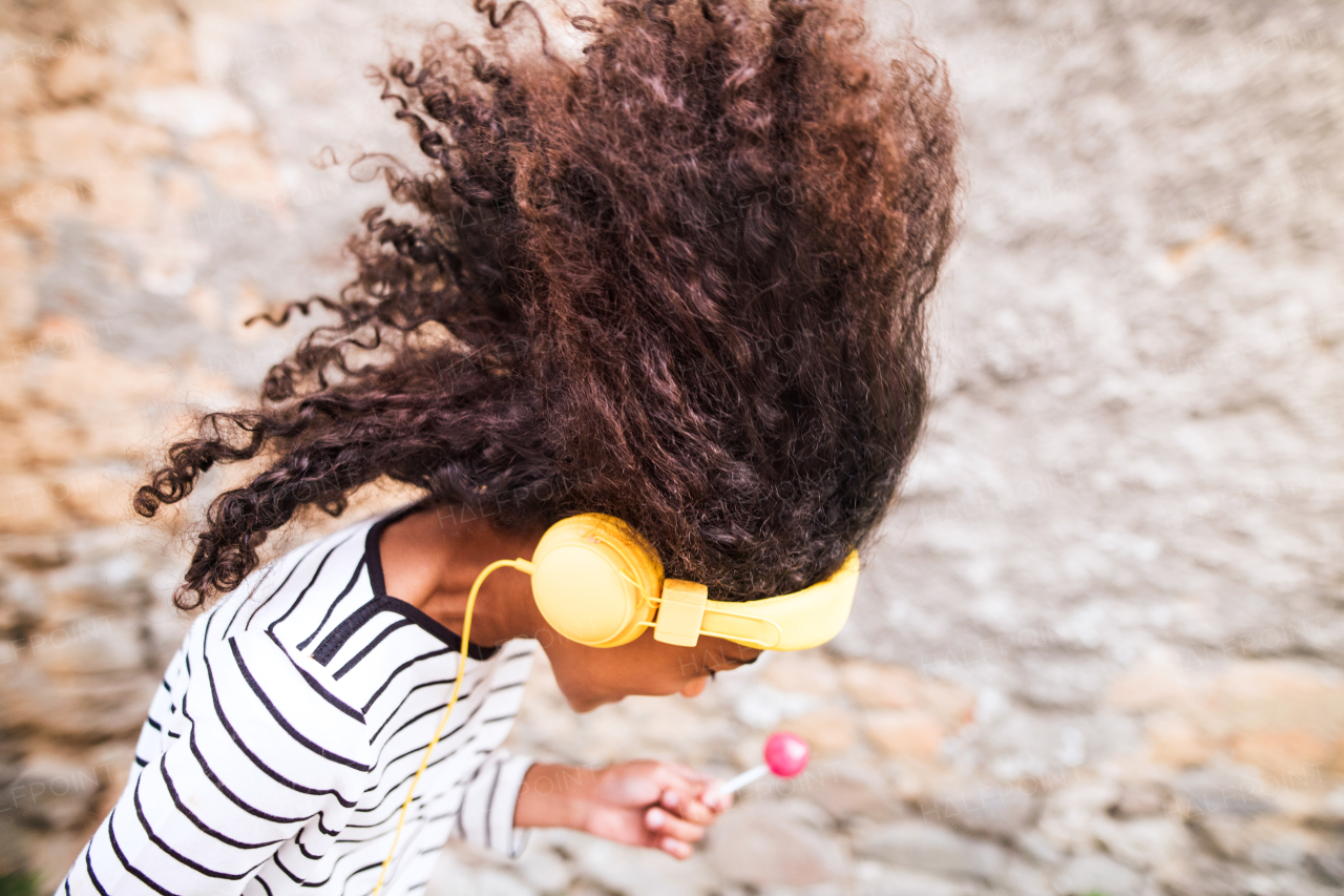 Beautiful african american girl with curly hair outdoors, against stone wall, wearing headphones, listening music, eating lollipop.