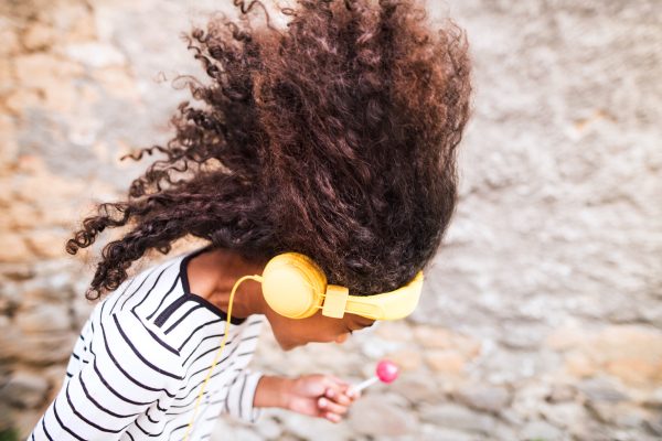 Beautiful african american girl with curly hair outdoors, against stone wall, wearing headphones, listening music, eating lollipop.