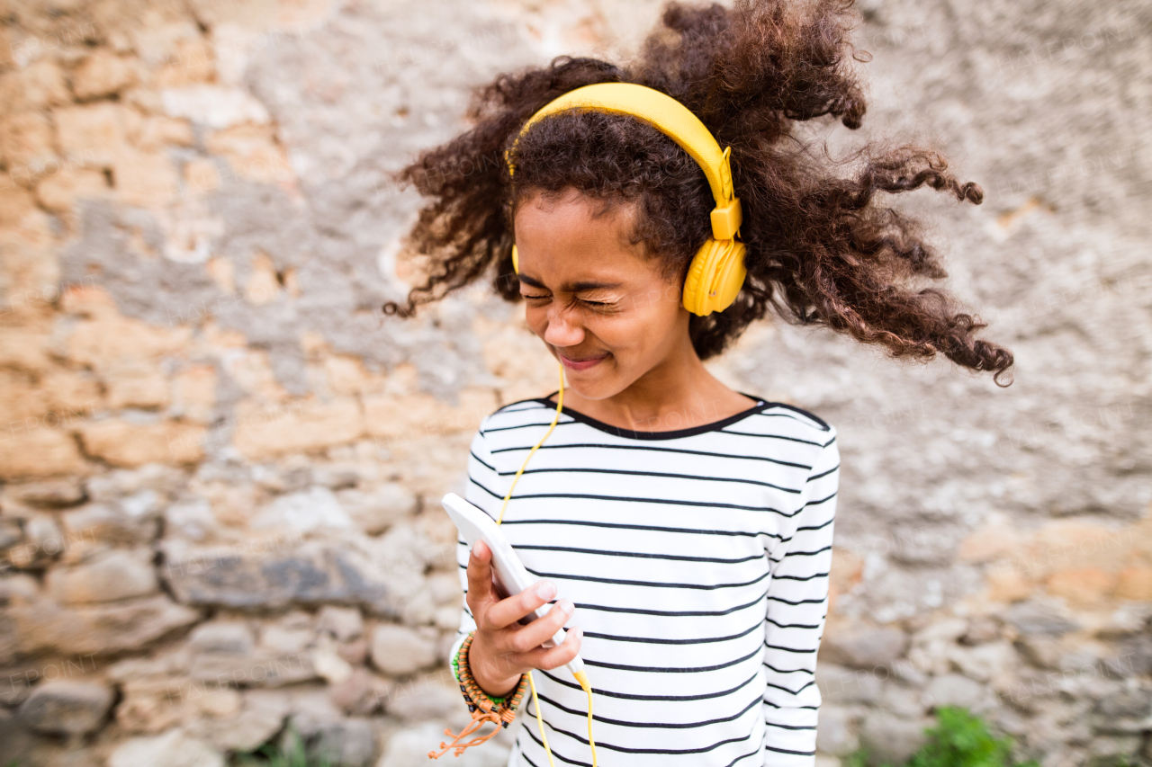 Beautiful african american girl with curly hair outdoors against stone wall, holding smart phone, wearing yellow headphones, listening music.