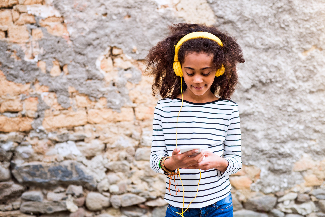 Beautiful african american girl with curly hair outdoors against stone wall, holding smart phone, wearing yellow headphones, listening music.