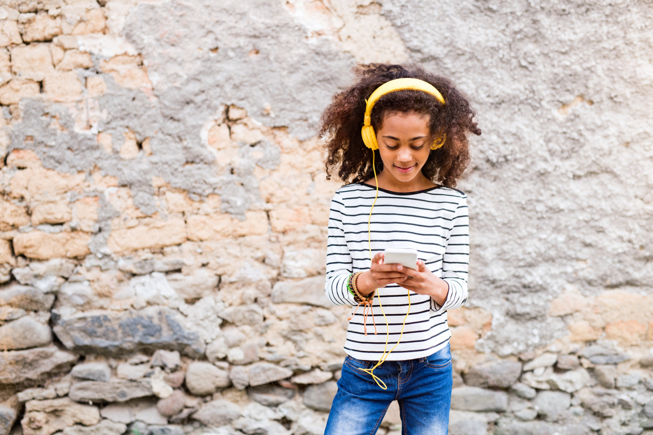 Beautiful african american girl with curly hair outdoors against stone wall, holding smart phone, wearing yellow headphones, listening music.