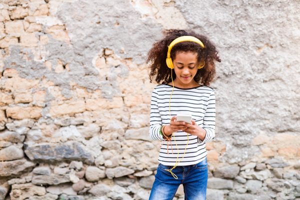 Beautiful african american girl with curly hair outdoors against stone wall, holding smart phone, wearing yellow headphones, listening music.