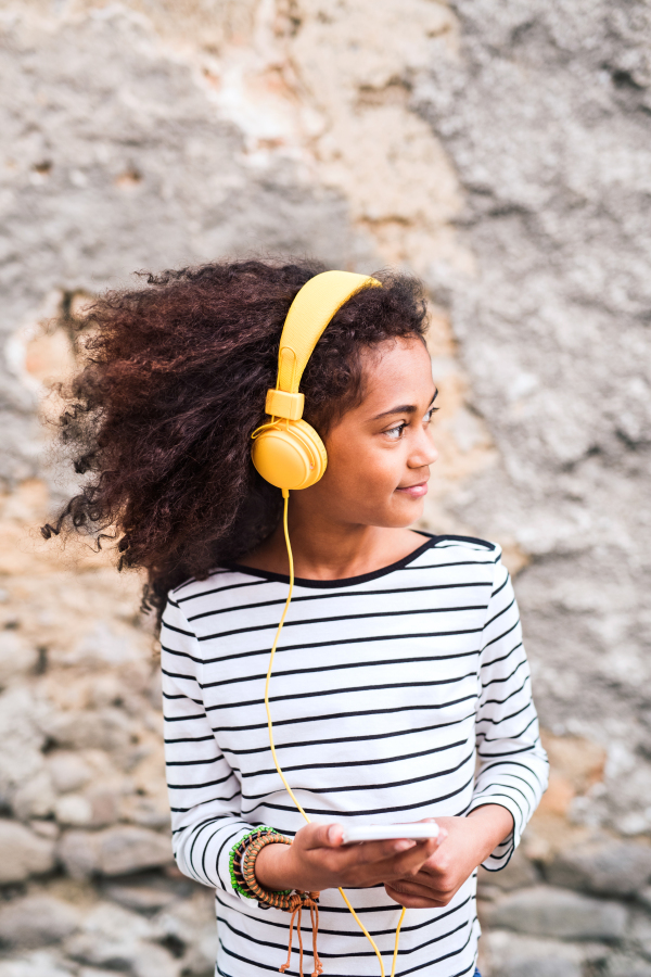 Beautiful african american girl with curly hair outdoors against stone wall, holding smart phone, wearing yellow headphones, listening music.