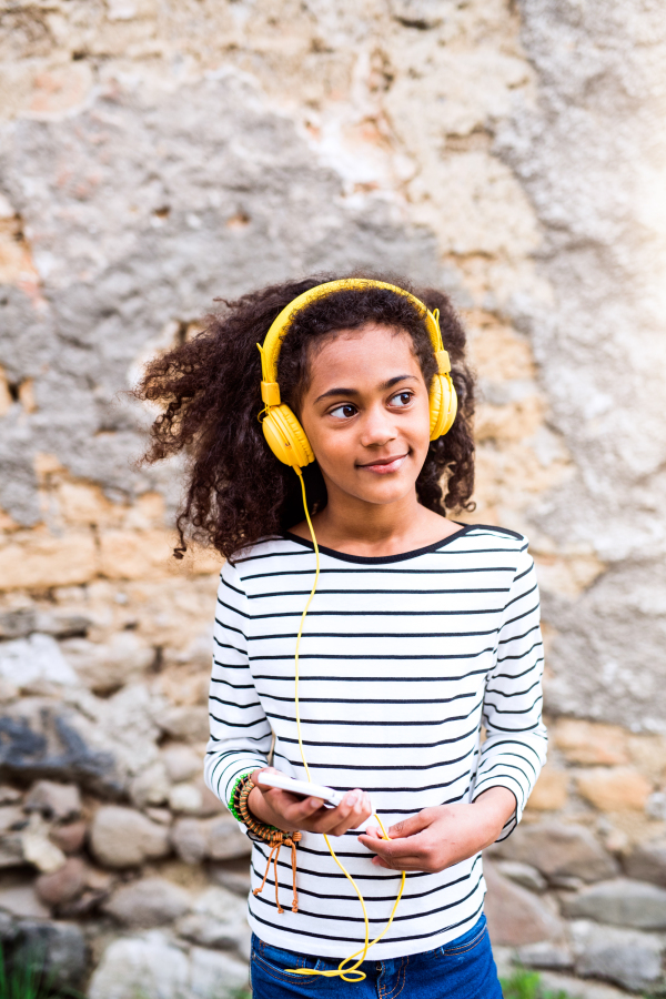 Beautiful african american girl with curly hair outdoors against stone wall, holding smart phone, wearing yellow headphones, listening music.