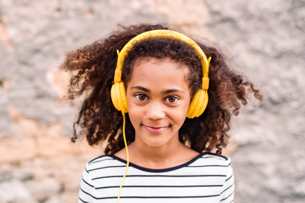 Beautiful african american girl with curly hair outdoors, against stone wall, wearing yellow headphones, listening music.