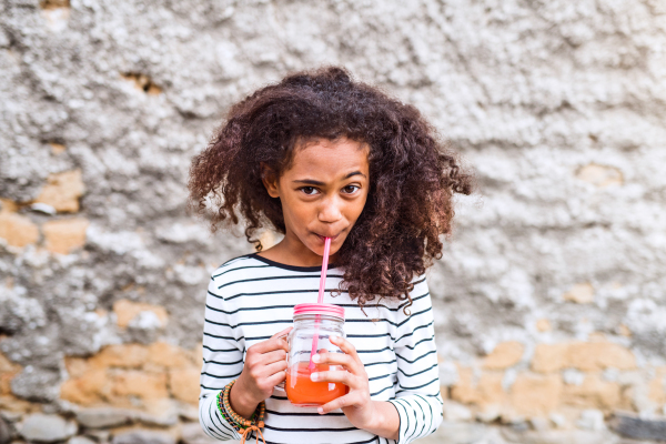 Beautiful african american girl with curly hair outdoors wearing striped t-shirt, drinking juice in glass with pink straw.