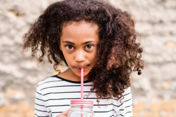 Beautiful african american girl with curly hair outdoors wearing striped t-shirt drinking water from glass with pink straw, standing against stone wall.