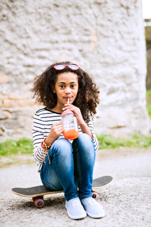 Beautiful african american girl with curly hair outdoors wearing striped t-shirt drinking juice from glass with pink straw, sitting on skateboard, stone wall background.