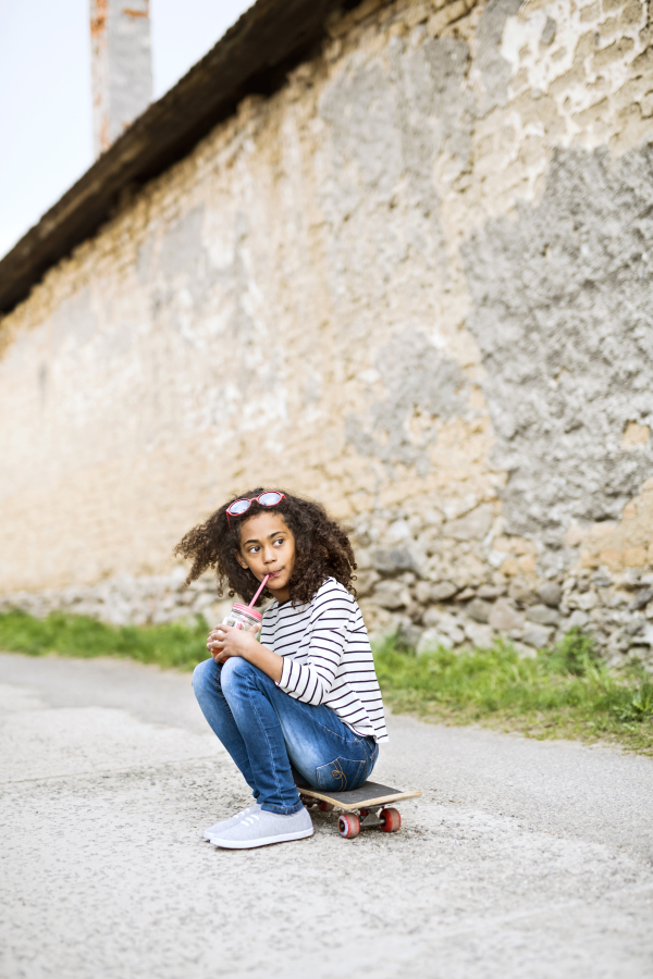 Beautiful african american girl with curly hair outdoors drinking juice from glass with pink straw, sitting on skateboard, stone wall behind her.