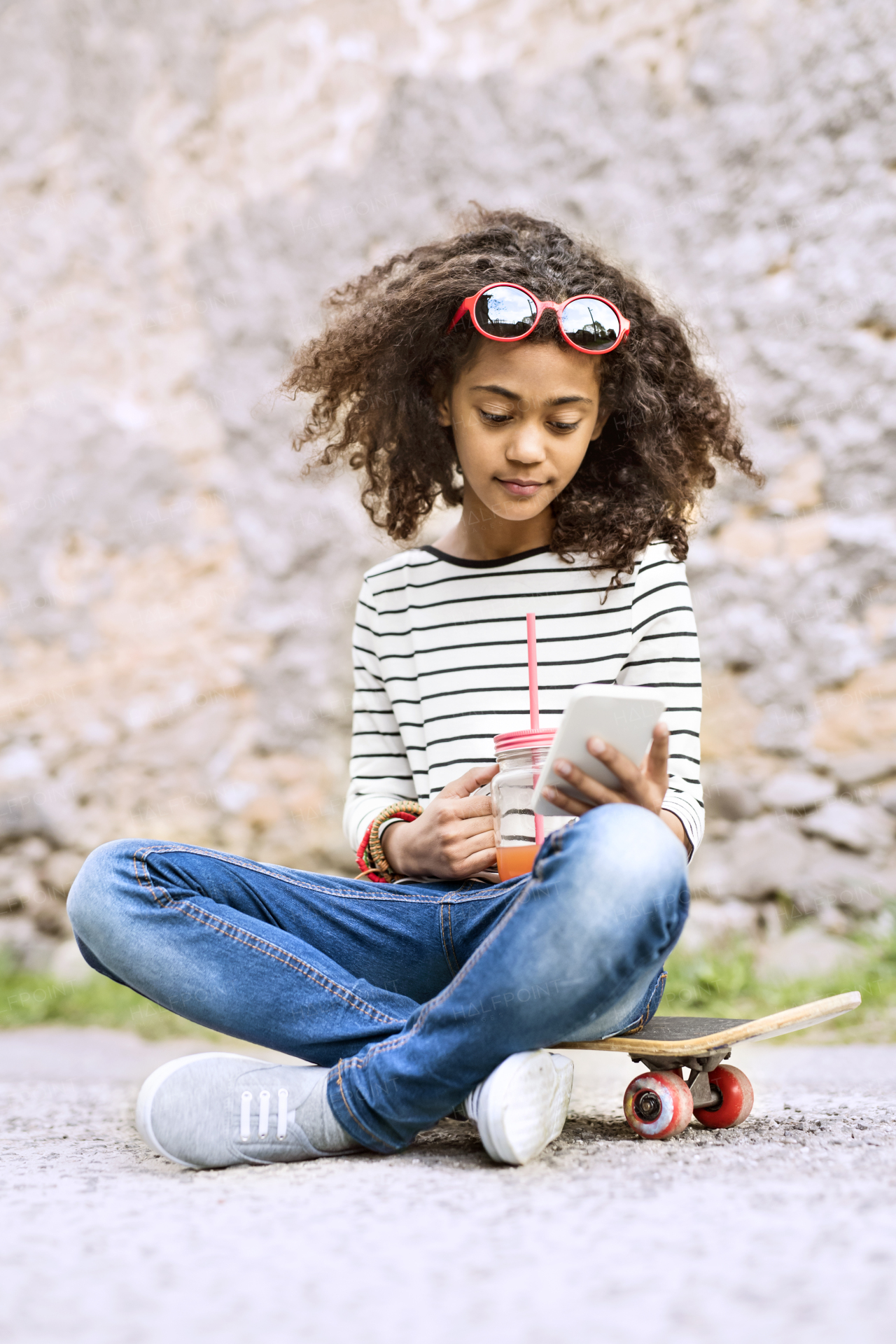 Beautiful african american girl with curly hair outdoors sitting on skateboard holding smart phone, watching something or texting, drinking juice in glass with pink straw.