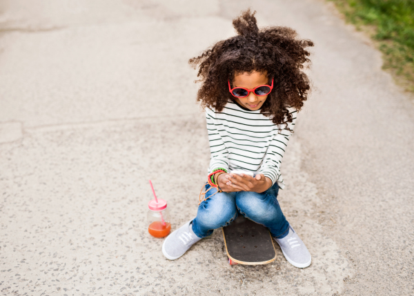 Beautiful african american girl with curly hair outdoors sitting on skateboard holding smart phone, watching something or texting, juice in glass with pink straw next to her.