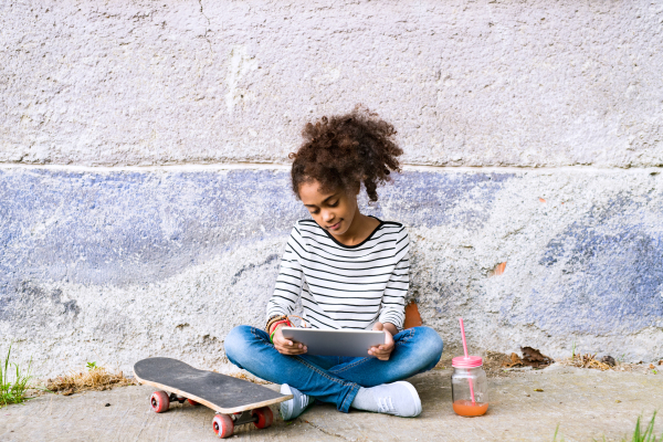 Beautiful african american girl with curly hair outdoors wearing striped t-shirt holding tablet, watching something, juice in glass with pink straw and skateboard laid on the ground next to her. Concrete wall background.