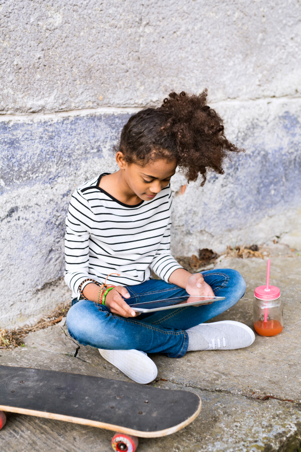 Beautiful african american girl with curly hair outdoors wearing striped t-shirt holding tablet, watching something, juice in glass with pink straw and skateboard laid on the ground next to her. Concrete wall background.