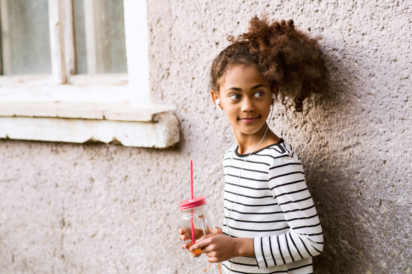 Beautiful african american girl with curly hair outdoors drinking juice from glass. Old house background.