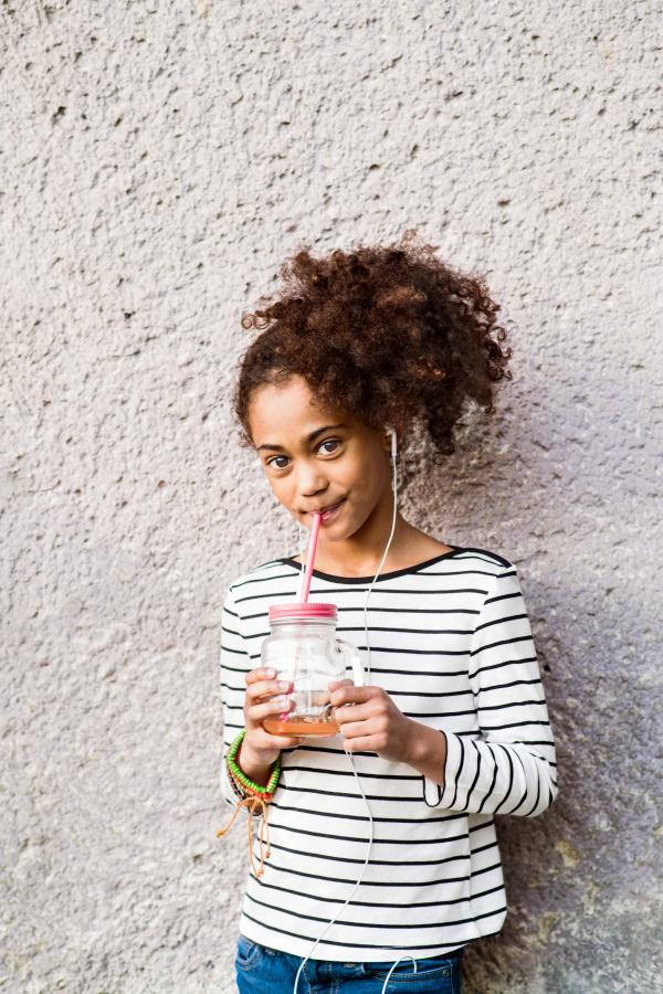 Beautiful african american girl with curly hair outdoors wearing striped t-shirt, earphones in her ears, listening music, drinking juice in glass with pink straw.