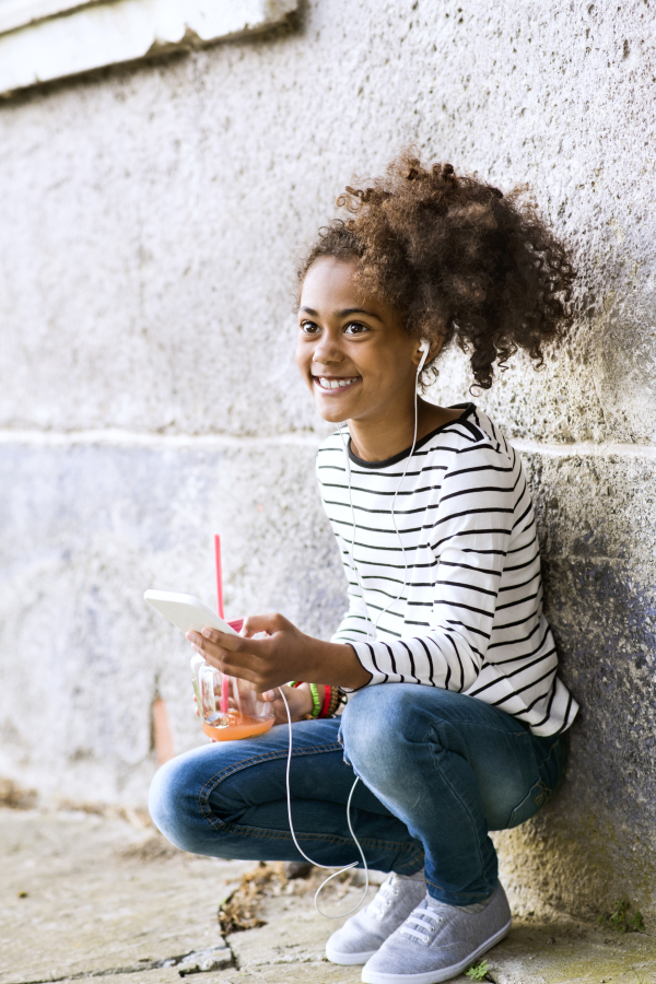 Beautiful african american girl with curly hair outdoors, holding smart phone, earphones in her ears, listening music, drinking juice from glass jar.