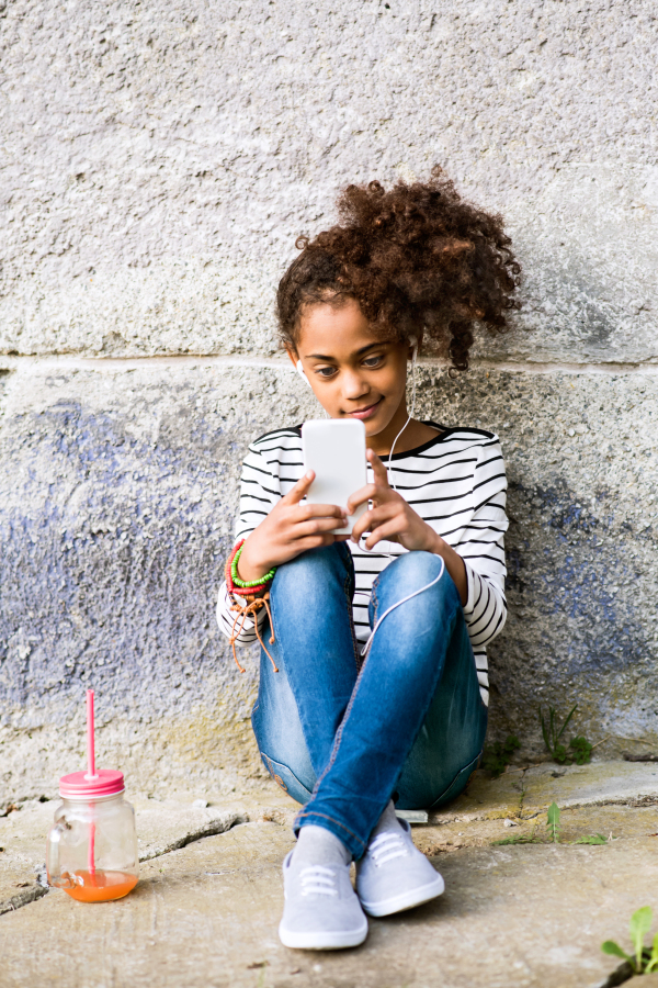 Beautiful african american girl with curly hair outdoors sitting on the ground, holding smart phone, earphones in her ears, listening music, juice in glass next to her.