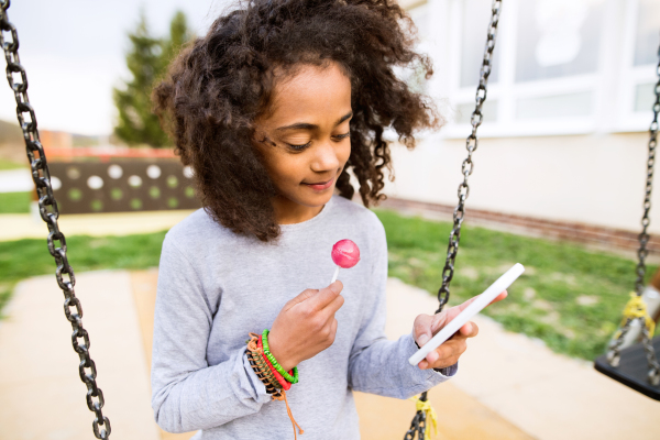 Beautiful african american girl on swing with smart phone writing text message or reading something, eating lollipop.