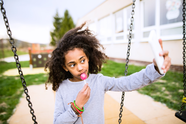 African american girl on swing eating lollipop taking selfie with smart phone.