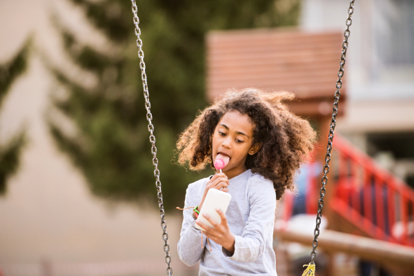 Beautiful african american girl on swing with smart phone writing text message or reading something, eating lollipop.