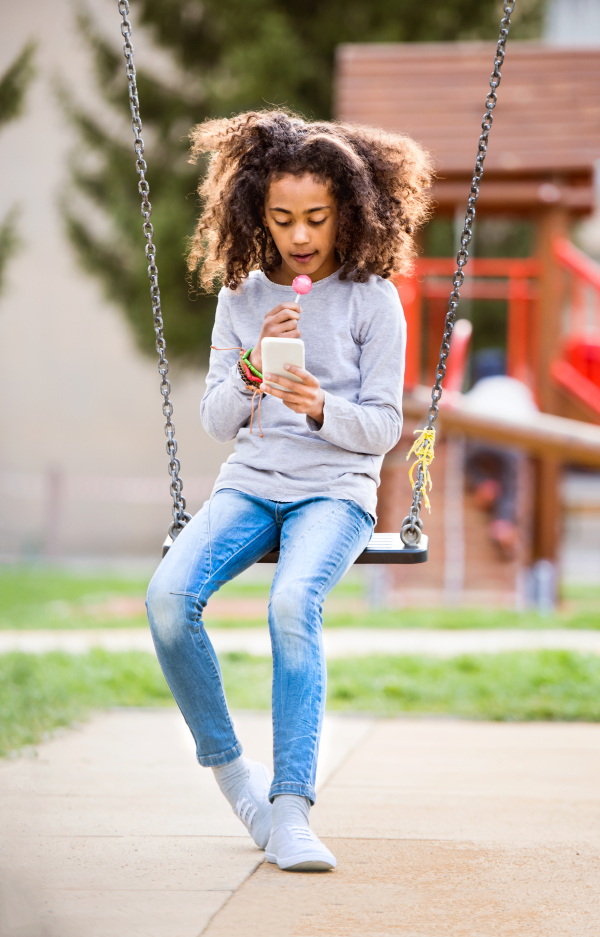 Beautiful african american girl on swing with smart phone writing text message or reading something, eating lollipop.