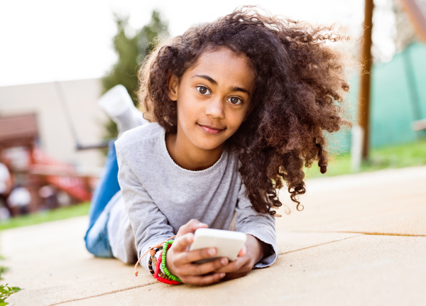 Beautiful african american girl with curly hair outdoors lying on the ground, holding smart phone, texting.