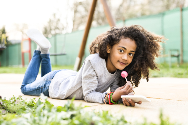 Beautiful african american girl with curly hair outdoors lying on the ground, holding smart phone, texting and eating lollipop.