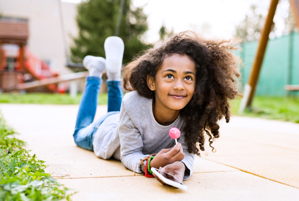 Beautiful african american girl with curly hair outdoors lying on the ground, holding smart phone, texting and eating lollipop.