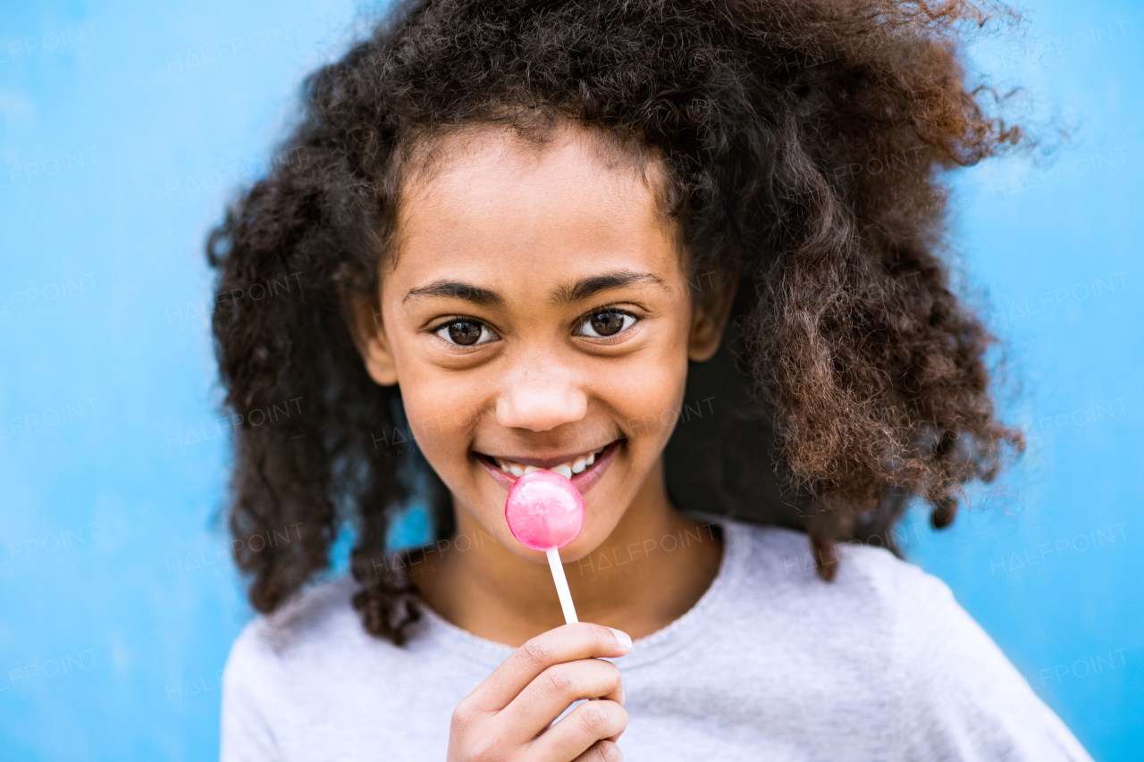 Beautiful african american girl with curly hair outdoors wearing gray sweatshirt, eating lollipop, standing against blue wall.