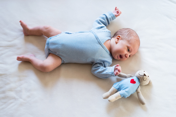 Cute newborn baby boy in blue striped onesie lying on bed, teddy bear toy with red heart next to him