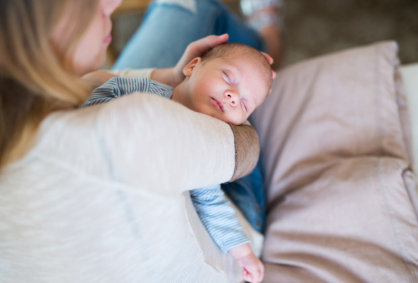 Unrecognizable young mother sitting on bed in bedroom holding her sleeping newborn baby son