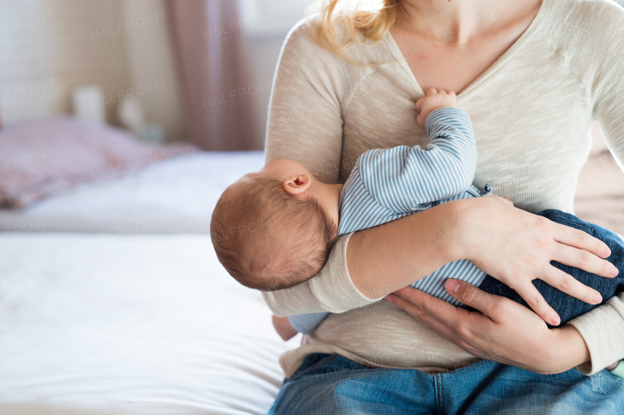 Unrecognizable young mother sitting on bed in bedroom holding her sleeping newborn baby son