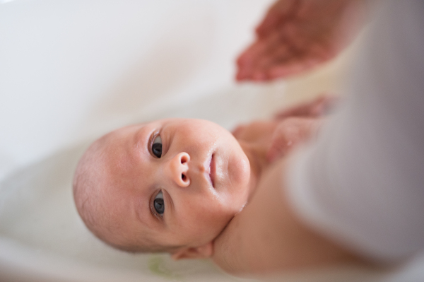 Unrecognizable mother holding her baby son, bathing him in small white plastic bath. Close up.