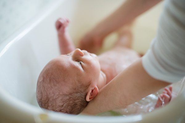 Unrecognizable mother holding her baby son, bathing him in small white plastic bath. Close up.