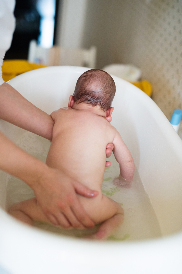 Unrecognizable mother holding her baby son, bathing him in small white plastic bath. Close up.