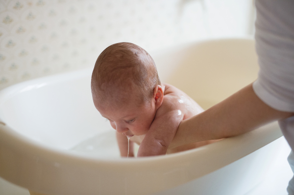 Unrecognizable mother holding her baby son, bathing him in small white plastic bath. Close up.