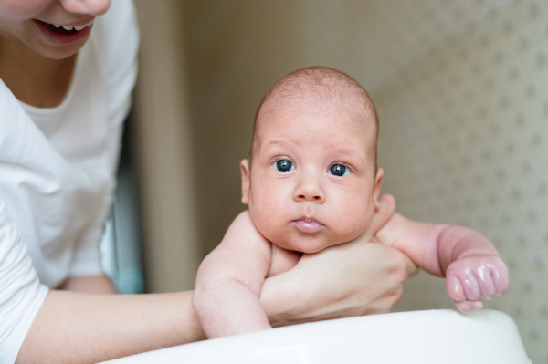 Unrecognizable mother holding her baby son, bathing him in small white plastic bath. Close up.