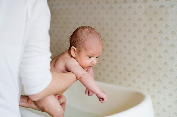 Unrecognizable mother holding her baby son, bathing him in small white plastic bath. Close up.