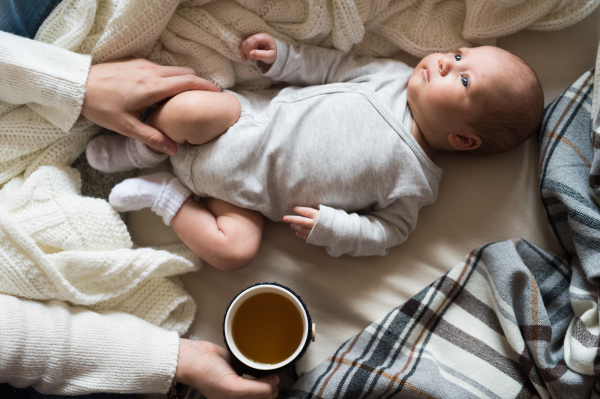 Unrecognizable mother holding a cup of tea and her newborn baby son lying on bed next to her