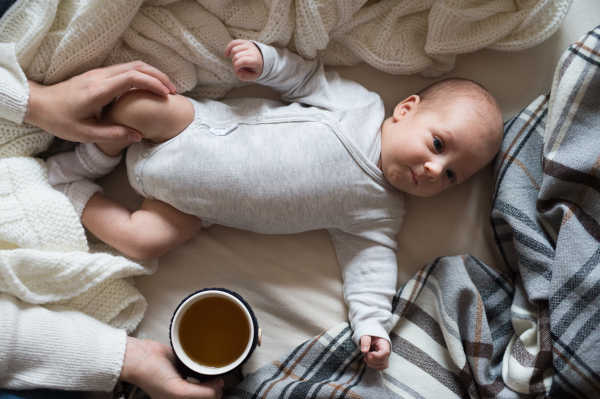 Unrecognizable mother holding a cup of tea and her newborn baby son lying on bed next to her