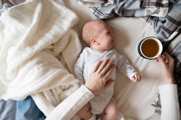 Unrecognizable mother holding a cup of tea and her newborn baby son lying on bed next to her