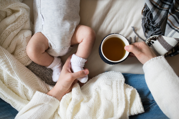 Unrecognizable mother holding a cup of tea and her newborn baby son lying on bed next to her