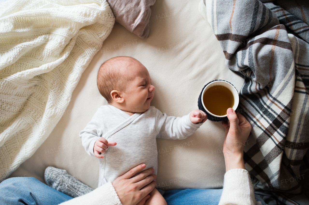 Unrecognizable mother holding a cup of tea and her newborn baby son lying on bed next to her