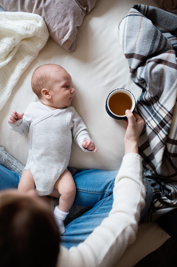 Unrecognizable mother holding a cup of tea and her newborn baby son lying on bed next to her