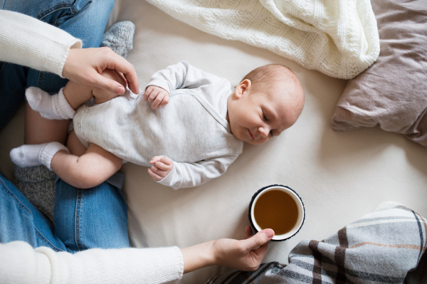 Unrecognizable mother holding a cup of tea and her newborn baby son lying on bed next to her