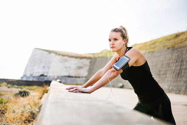 Young sporty woman runner with earphones and smartphone in armband standing outside on the beach in nature, listening to music and resting.