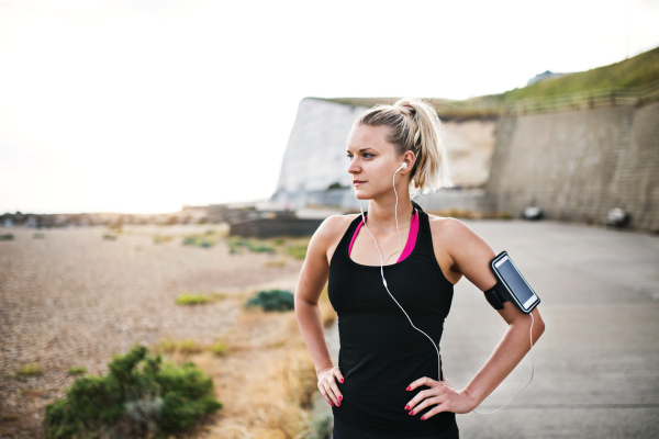 Young sporty woman runner with earphones and smartphone in armband standing outside on the beach in nature, listening to music and resting.